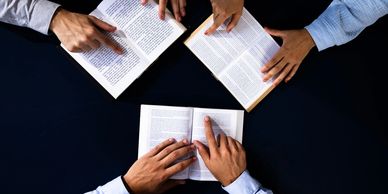 Three people pointing at text in open books on a dark table.