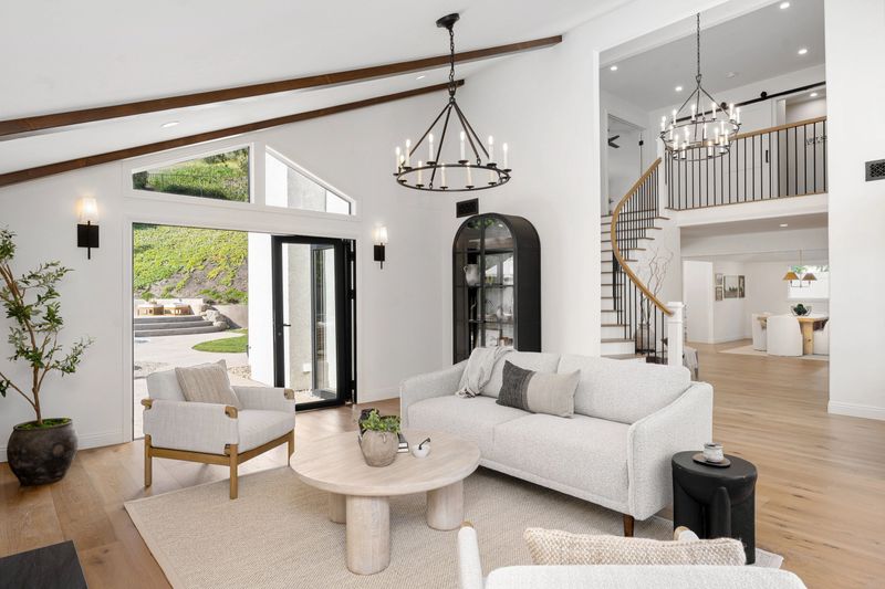 A modern living room with white furniture, wooden floor, and large windows overlooking a backyard.