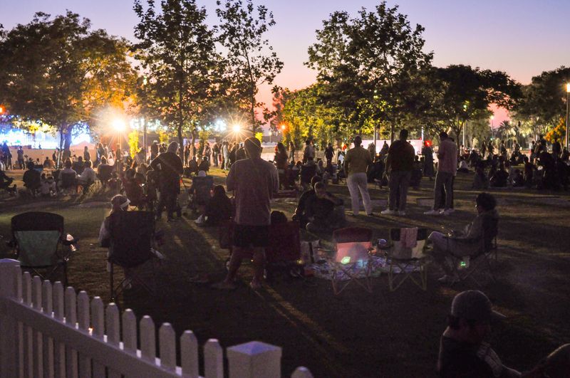 This image showcases a vibrant community event taking place at sunset. People of all ages are gathered in a park, enjoying an evening outdoors. The warm glow of the setting sun, coupled with the artificial lights, creates a festive atmosphere. Families and friends are seated on the grass, chatting, and relaxing, embodying the spirit of togetherness and community.