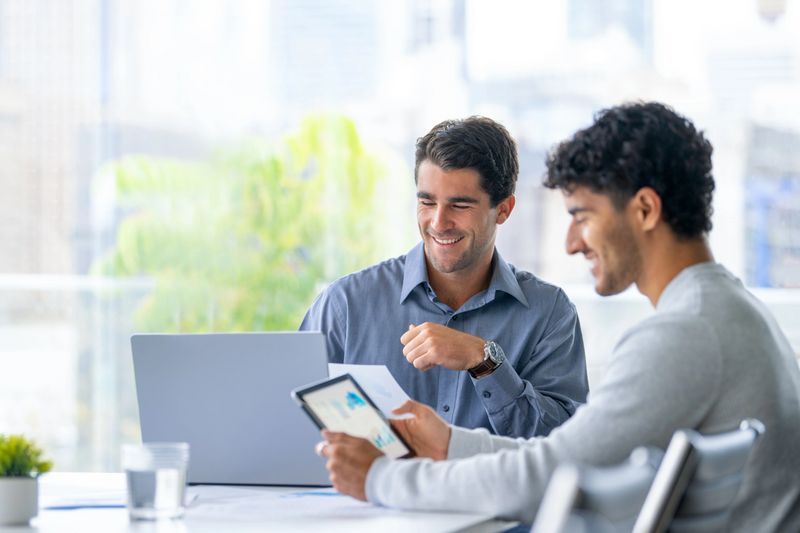 Two businessmen working on a digital tablet and laptop computer in the office. Finance Charts and graphs can be seen on the digital tablet