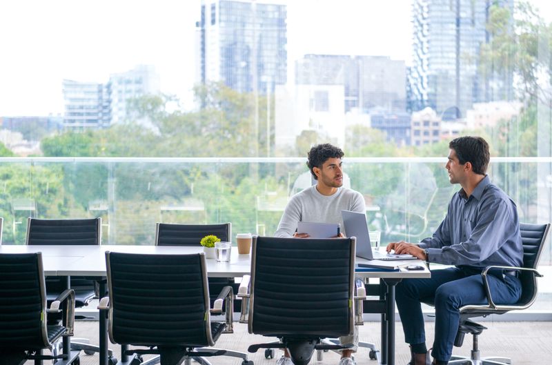 Two businessmen working on a digital tablet and laptop computer in the office a board room or meeting room. There is a large window behind them