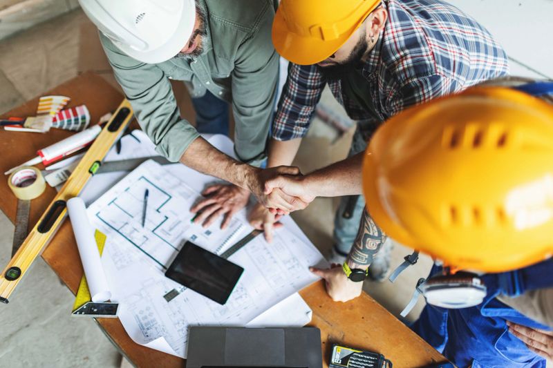 Home owner shaking hands with construction worker with blueprints and working tools at background. Contract between foreman and constraint workers team. Handshake. High angle view.