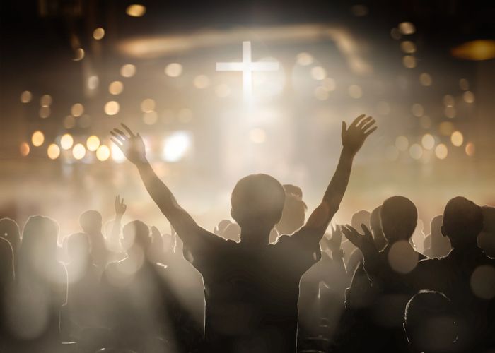Silhouetted worshippers with raised hands in a church illuminated by a glowing cross.
