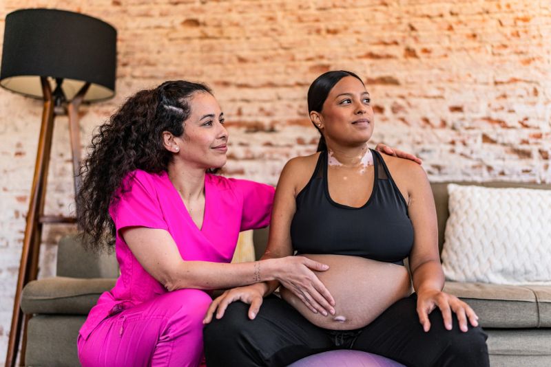 Young pregnant doing physical therapy using ball with physiotherapist at house call
