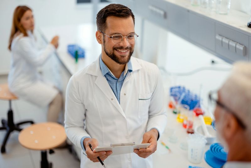 Modern Medical Research Laboratory: Young Male Scientist Holding Clipboard and Talking to His Faceless Mature Male Colleague in a Modern Laboratory