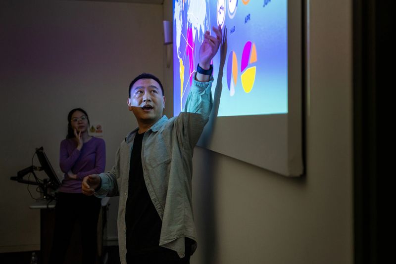 A medium shot of a university lecturer pointing at a projector screen with an open palm. He is looking at the screen with an expression of concentration and wearing smart casual clothing. There is a colleague in the background waiting patiently to change to the next slide on the computer. The projector screen shows a presentation about data mining at a university campus in London.

Videos are available similar to this scenario.