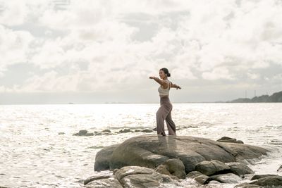 A woman practices yoga on a rock by the ocean under a cloudy sky.