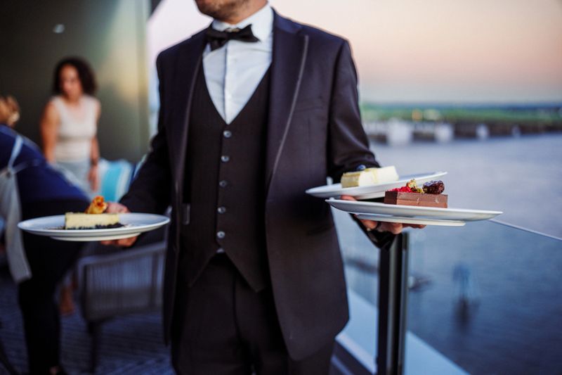 Cropped view of an unrecognizable male restaurant server, with selective focus on his hands, as he carries three desserts to a table of patrons dining on the rooftop terrace at sunset.