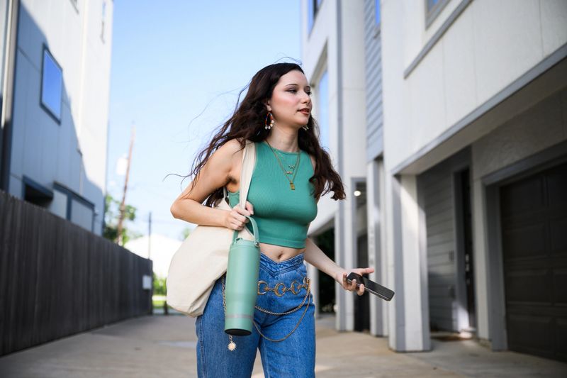 Low angle view of generation z woman in jeans and turquise crop top wearing gold jewelry walking down road between apartment blocks carrying mobile phone tote bag and water bottle