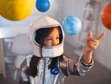 A child in a silver astronaut costume pointing at hanging planets.