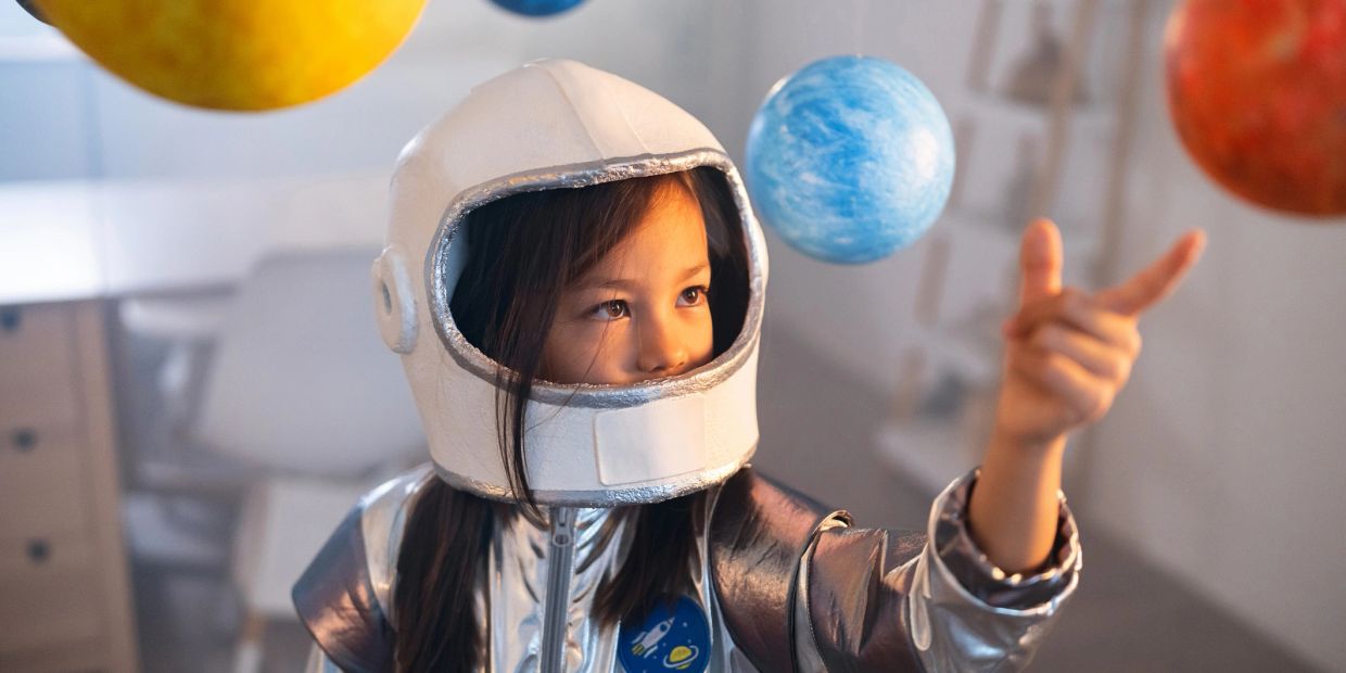 Child in an astronaut costume pointing at floating planets.
