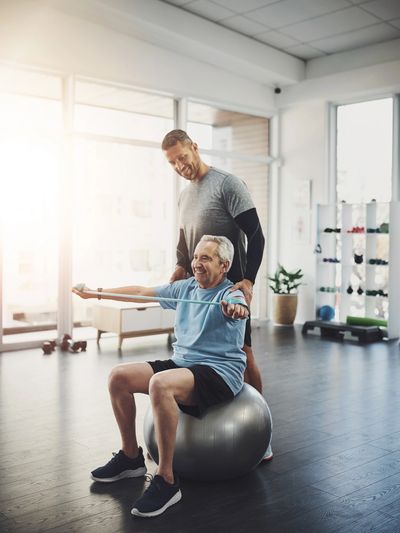 Senior man exercising with resistance band on stability ball, guided by a trainer.