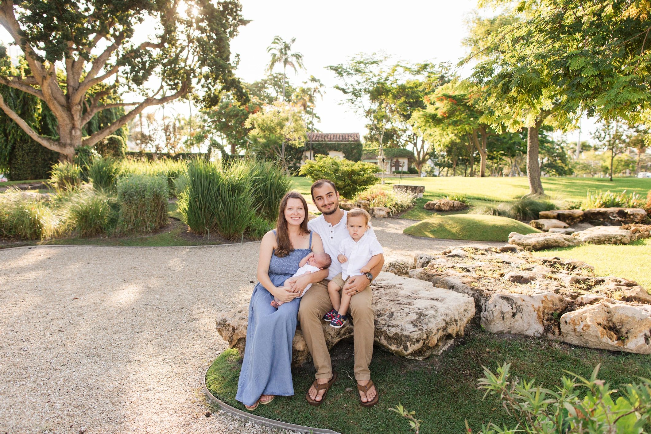 Happy family of four posing in a sunny park with lush greenery.