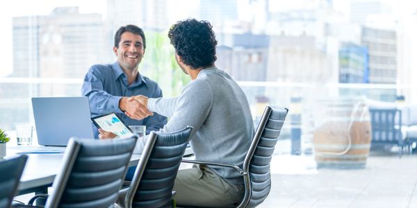 Two men shaking hands in a bright office with laptops and charts.