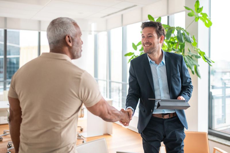 A smiling elderly Hispanic man shakes hands with a financial advisor in an office, showcasing a professional interaction with a diverse clientele.