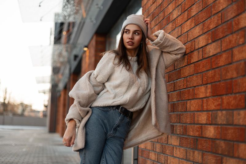 Fashionable young woman model in a stylish hat in a trendy coat in blue jeans in a vintage sweater is resting near a vintage brick wall on a street in the city. Attractive urban girl. Spring style.