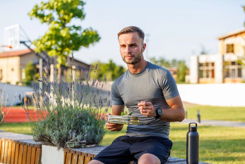Confident young man sitting in an outdoors gym and eating healthy salad after hard workout.