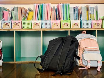 Two backpacks on the floor in front of colorful magazine holders on a shelf.