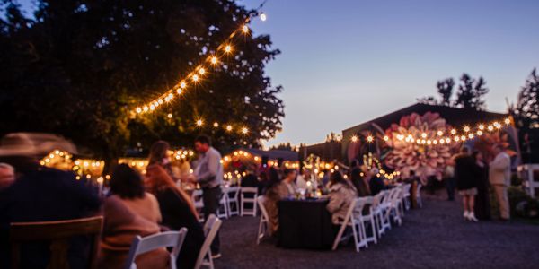 Outdoor evening gathering with string lights and people socializing at tables.