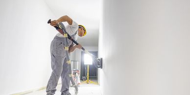 Worker sanding a white wall in a bright, empty hallway.