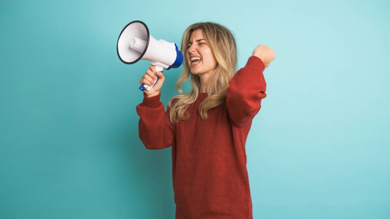Young female activist in maroon pullover with blond hair shouting on megaphone while protesting against blue background