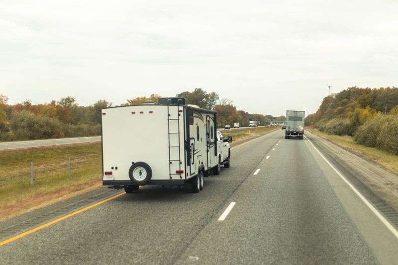 A white truck towing a trailer on a rural highway during the day, representing the freedom of road travel and the utility of recreational vehicles.