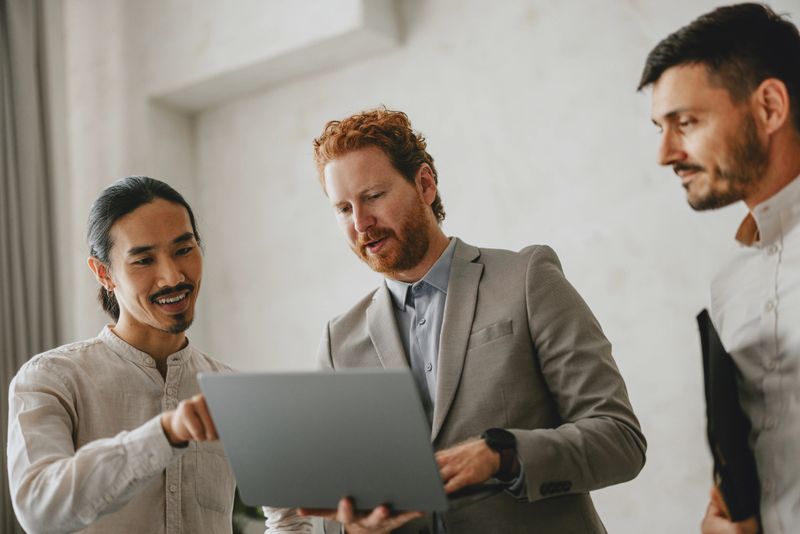 Three professionals in a business meeting at a bright beige office, discussing work on a laptop. Collaborative and productive atmosphere.