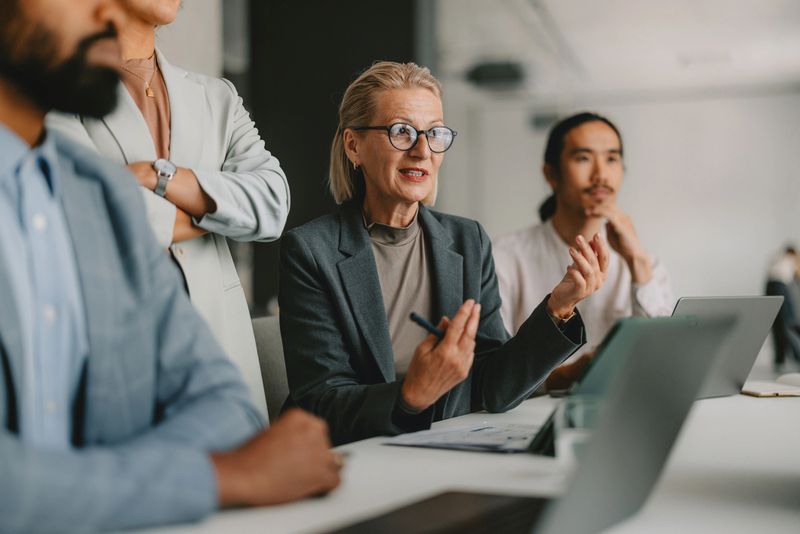 A diverse group of professional individuals engaged in a business meeting within a bright beige office, illustrating teamwork, collaboration, and leadership.
