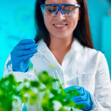 Scientist in lab coat and gloves handling a pipette near green plants.