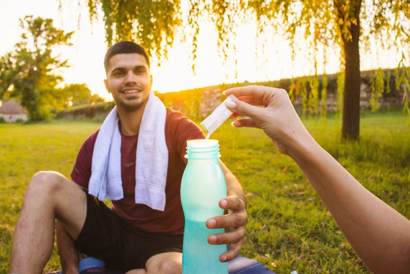 Unrecognizable female Caucasian athletes pouring powdered health supplements into a reusable water bottle, after she finished an outdoor exercise with an boyfriend or male friend