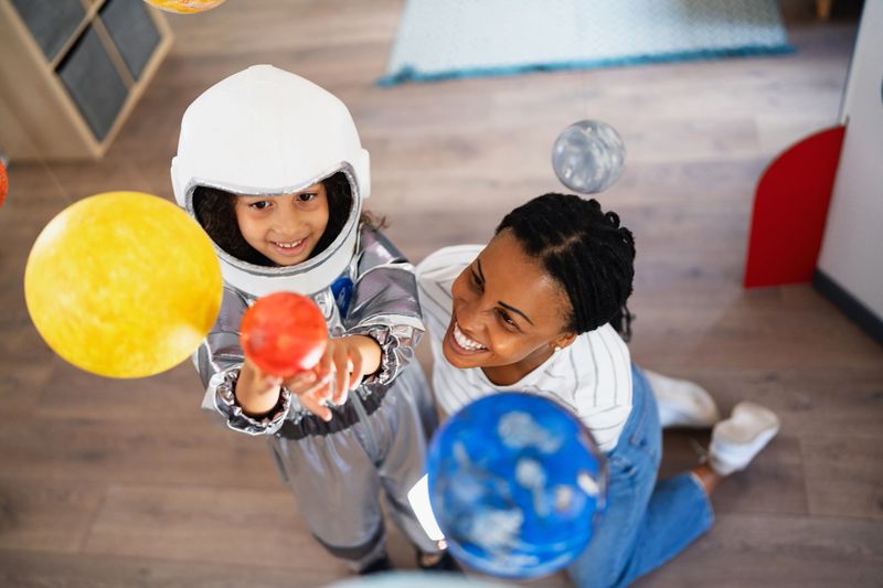 Mid-adult mother of Black ethnicity, playing with her daughter while she wear a space suit, and exploring handmade planets