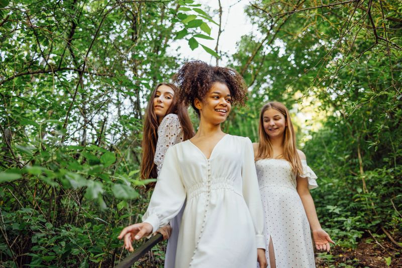 Three happy pretty smiling teenage girls in white summer elegant dresses walk along path in forest or park. Cheerful beautiful girls have a good time together. Green trees on background. Friendship.