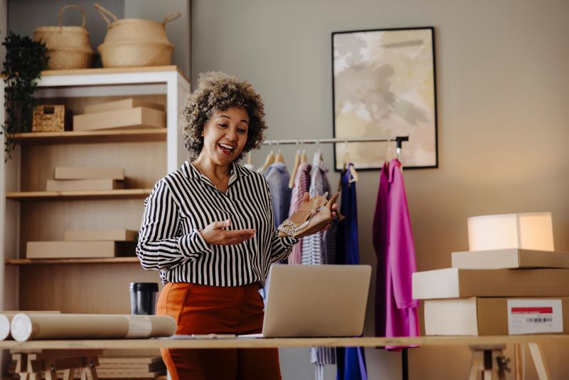 Female small business owner smiling cheerfully while standing in her shop. Happy businesswoman managing her clothing orders on a laptop. Black female entrepreneur running an online clothing store.