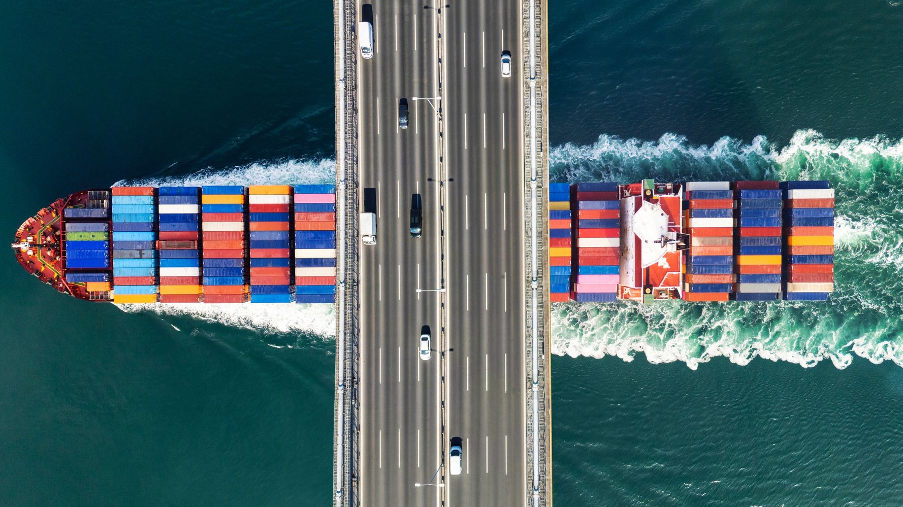 A cargo ship passes under a bridge over the sea, carrying colorful containers.