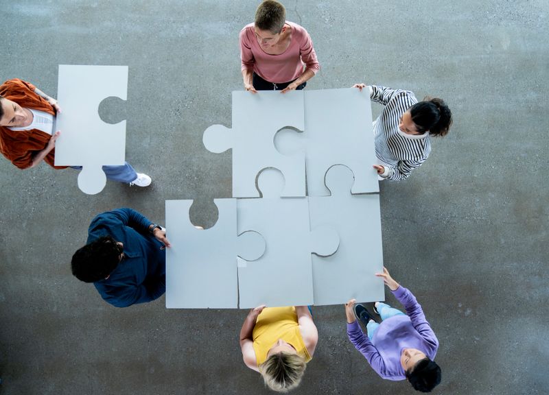 A small group of adults are seen each holding a large puzzle piece as they work on a teambuilding exercise at work.  They are each dressed casually and are focused on working together to solve the problem in this aerial view.