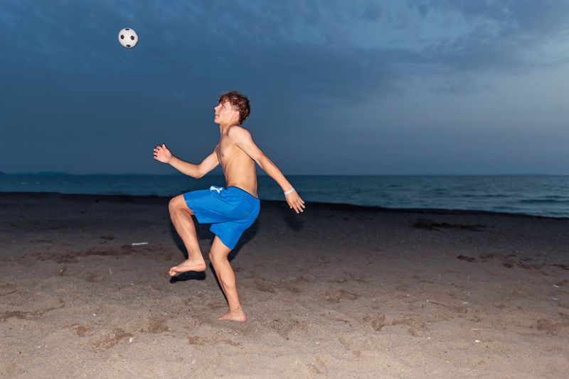 Teenage boy playing soccer on a sandy beach. The boy is jumping to perform a bicycle kick.Summer vacations day evening in Tuscany, Italy. Retro styled photo with camera flash used.Shot with Canon R5