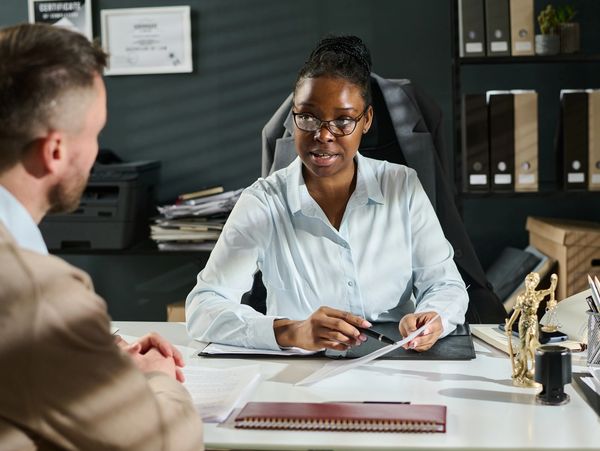 A woman in glasses talks to a man across a desk in an office.