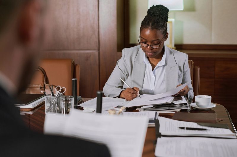 Young serious female notary in formalwear signing or certifying paper documents after reading them while sitting in boardroom
