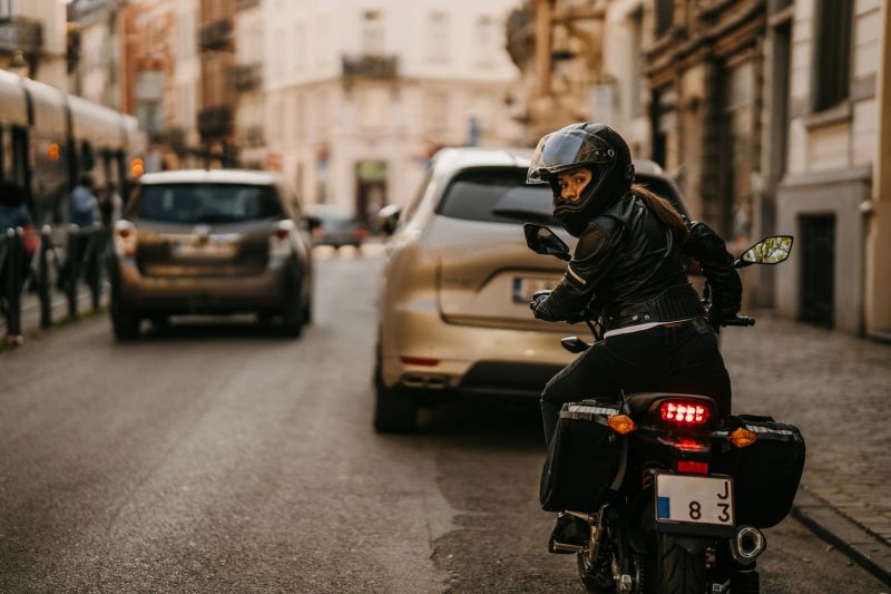 Rear view shot of female rider on motorcycle is driving her motorbike, wearing helmet, leather jacket and gloves in the city