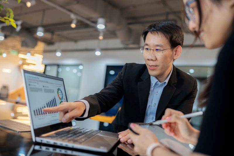 A financial advisor offers expert advice on financial planning and asset management to clients in a modern office setting. The advisor points at a graph on the computer screen, explaining investment strategies and financial management