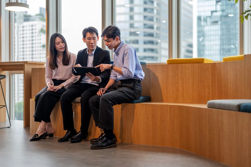 A financial planner sits in a contemporary coworking space with a backdrop of city skyscrapers, discussing investment strategies with clients. The advisor uses a digital tablet to analyze financial data, providing tailored advice on savings, banking, and investment options.