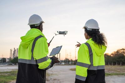 Two workers in high-visibility jackets operate a drone outdoors at sunset.