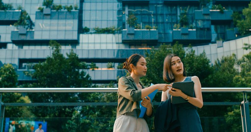 Two young Asian businesswomen meeting business strategy discussion talk outside modern office building. Sustainable business outdoor concept.
