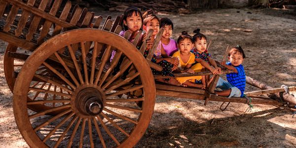 Six children sitting on a traditional wooden cart outdoors in a rural setting.