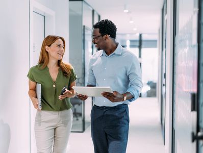 Two colleagues walking and talking in a modern office hallway.