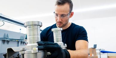 A man in safety glasses and gloves working on metal piping in a workshop.