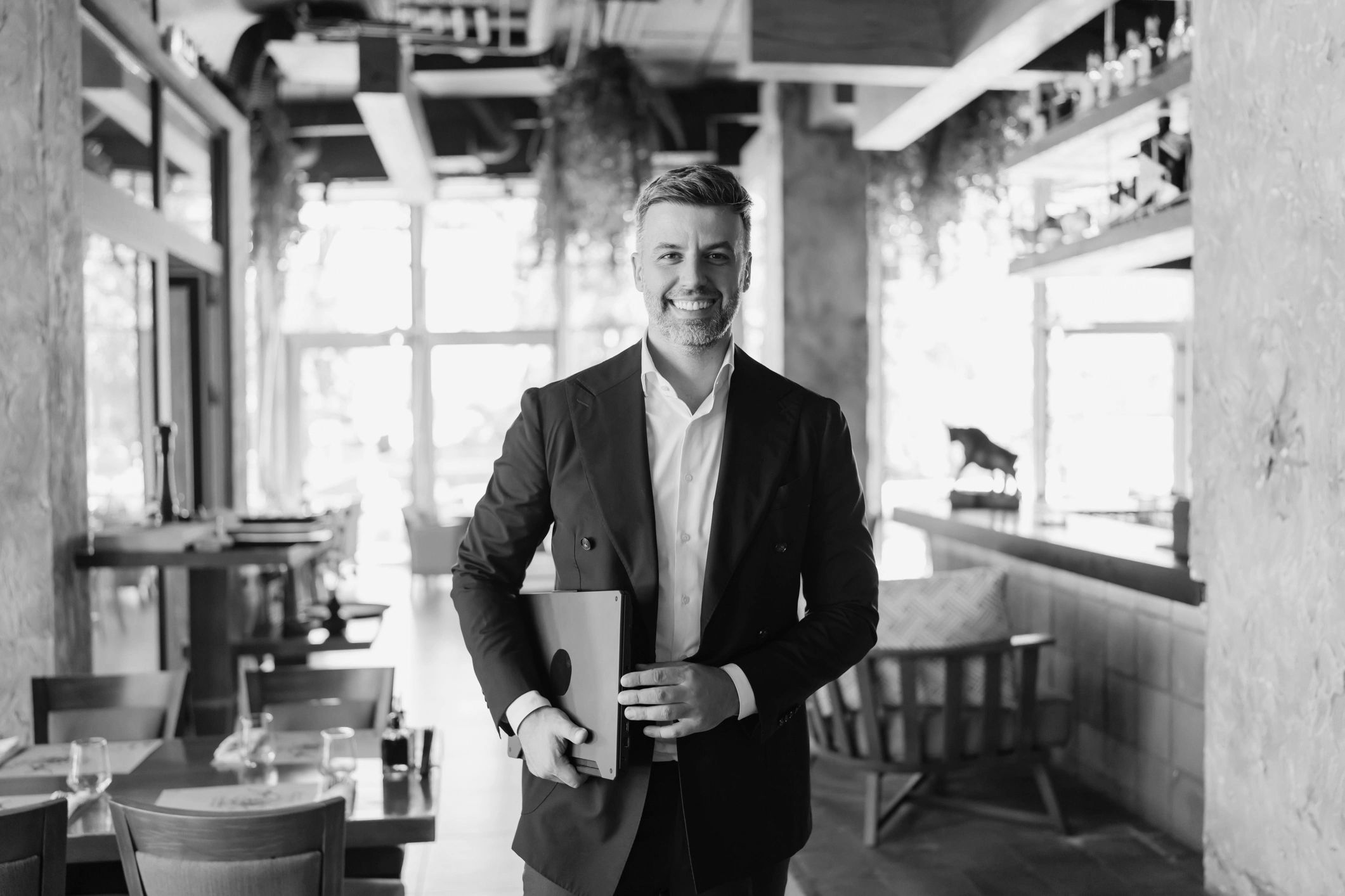 Smiling businessman in a suit holding a laptop in a modern cafe.