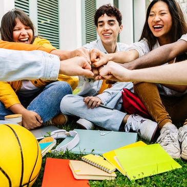 A diverse group of friends sitting on grass, fist-bumping and smiling together.