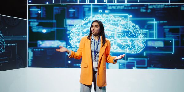 A woman in an orange blazer speaks in front of a digital brain projection.