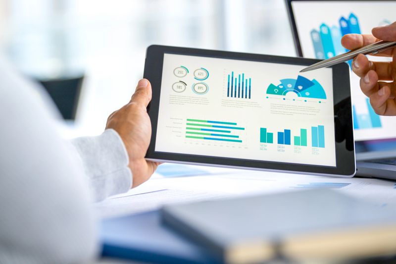 Two business men meeting and looking at financial data, charts and graphs on a digital tablet and a laptop computer. There are documents and paperwork on the table. One man is pointing at the screen
